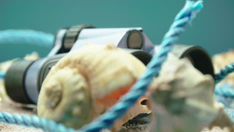 Close-up-product-video-of-blue-and-black-binoculars-with-sea-shells-and-blue-rope,-surrounded-by-beach-sand,-on-a-turntable-display