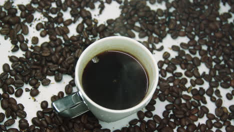 Weiße-Heiße-Tasse-Kaffee-Mit-Dampf,-Umgeben-Von-Gerösteten-Kaffeebohnen,-Die-Sich-Auf-Einem-Drehteller-Drehen