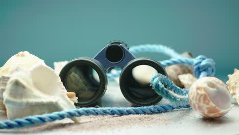 Blue-and-black-binoculars-with-sea-shells-and-blue-rope,-surrounded-by-beach-sand,-on-a-turntable-display