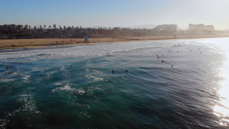 Huntington-Beach,-California-surf-at-sunrise-from-4k-drone,-wide-static,-with-waves,-beach,-and-hotels,-with-group-of-surfers-waiting-to-surf