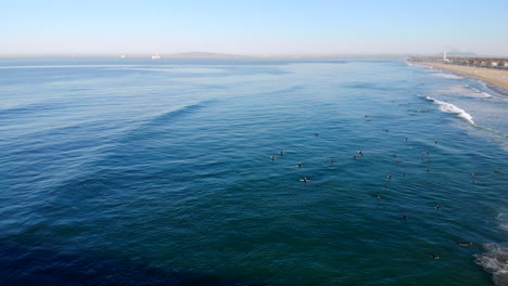 Wide-aerial-4k-HD-drone-push,-flyover-Huntington-Beach-Pier-to-large-group-of-surfers-in-sunny,-blue-Pacific-Ocean,-Southern-California-early-morning