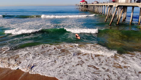 Beautiful-aerial-4k-drone-pulls-back-at-Huntington-Beach-Pier-surf-to-reveal-female-surfer-girl-paddling-out-through-waves-in-early-morning-Pacific-Ocean,-Southern-California