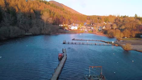 Aerial-crane-down-shot-of-a-harbour-at-the-Highlands-of-Scotland-during-golden-hour,-showing-Balmaha
