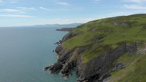 Drone-flying-backwards-in-direction-of-the-sea-from-Beautiful-cliffs-on-a-green-rocky-mountain-by-the-Irish-blue-sea