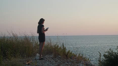 Young-woman-standing-on-a-cliff-by-the-sea-on-sunset,-takes-a-picture-with-tablet-computer-,-wide-shot