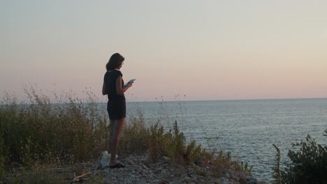 Young-woman-standing-on-a-cliff-by-the-sea-on-sunset,-with-tablet-computer-in-hands,-wide-shot