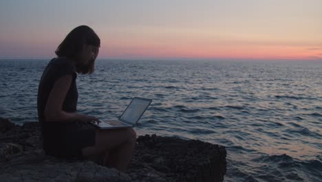 Mädchen-Mit-Laptop-Sitzt-Auf-Felsen-Am-Meer-Bei-Sonnenuntergang,-Zeitlupe