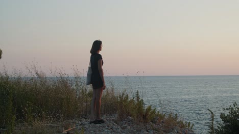 Girl-standing-on-a-cliff-by-the-sea-on-sunset--golden-hour,-wide-shot