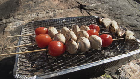 Skewers-with-cherry-tomatoes-and-mushrooms-cooking-on-a-disposable-BBQ-grill-on-the-rocks-by-the-ocean-side