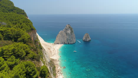 Aerial-view-reveal-from-woodland-cliff-edge-to-yacht-sailing-in-Zakynthos-turquoise-ocean-island-travel-destination-tilt-down-shot