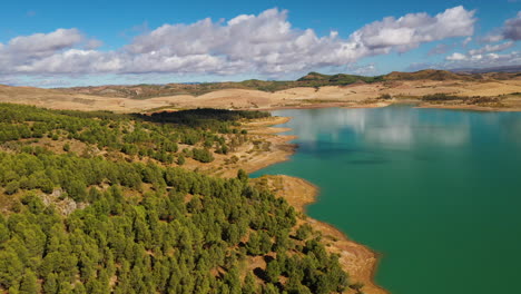 Panning-aerial-drone-shot-of-a-cyan-coloured-lake-surrounded-by-a-rocky-dessert-landscapes-and-forest