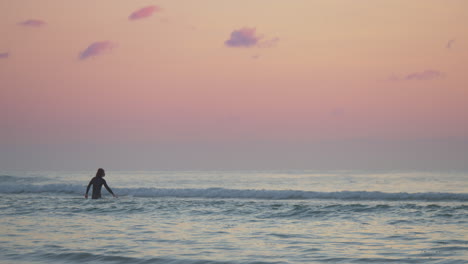 Männlicher-Surfer-In-Silhouette,-Der-Bei-Sonnenuntergang-Im-Meer-Gegenüber-Den-Wellen-Spazieren-Geht