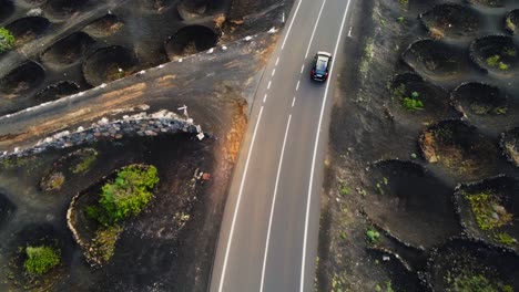 Vehicle-driving-through-asphalt-road-surrounded-by-holes-with-winery-plants-in-Lanzarote,-aerial-drone-view