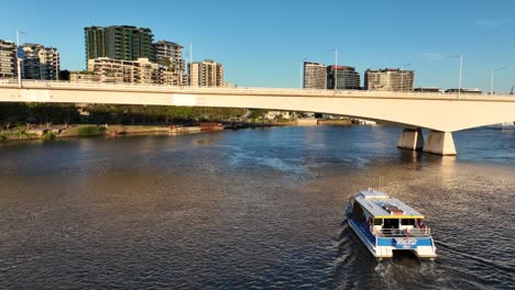 Drone-shot-tracking-City-Cat-Boat-on-Brisbane-River-as-we-pass-underneath-M3-Expressway-bridge,-with-South-Bank-and-Brisbane-CBD-in-background