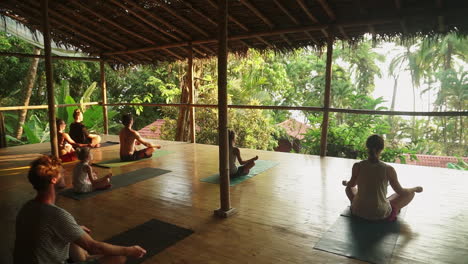 Un-Grupo-De-Viajeros-Sentados-En-Meditación-Durante-La-Puesta-De-Sol-En-Un-Soleado-Porche-De-Madera-Con-Vistas-A-La-Selva-Tropical-Y-La-Selva-Que-Conduce-A-La-Playa