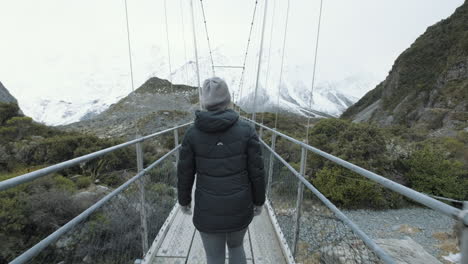 Eine-Folgende-Aufnahme-Von-Hinten-Einer-Frau,-Die-An-Einem-Kalten-Wintertag-In-Neuseeland-über-Eine-Drehbrücke-über-Einen-Fließenden-Bach-Geht,-Der-Von-Schneebedeckten-Bergen-Umgeben-Ist