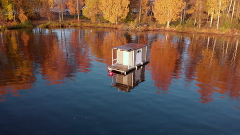 Point-of-view-aerial-of-a-small-white-houseboat-in-a-beautiful-lake-close-to-a-colorful-forest-in-the-autumn