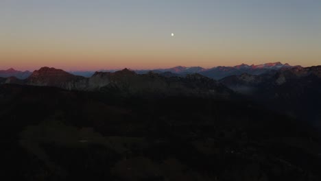 Aerial-shot-of-Mountain-range-at-sunset,-pink-summits-and-the-moon-rising-The-Alps,-Switzerland