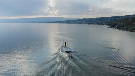 Toma-Aérea-Siguiendo-Un-Barco-De-Vapor-Belle-epoque-En-El-Lago-Léman-Frente-A-Cully,-Lavaux---Suiza-Agua-Similar-A-Un-Espejo-Y-Nubes-De-Tormenta-En-El-Fondo