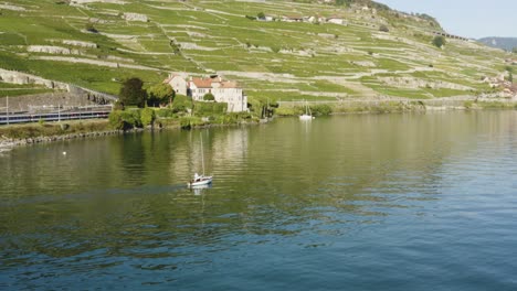 Aerial-shot-of-sailboat-in-front-of-castle-on-the-shore-of-Lake-Léman-in-Lavaux-vineyard,-plus-Swiss-train-passing-just-behind-the-castle-Chateau-de-Glérolles---Lavaux,-Switzerland