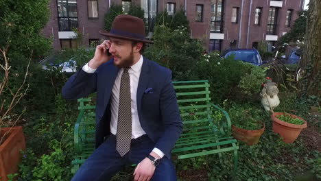 Un-Hombre-Elegante-En-Estilo-Dandy-Con-Traje-Azul-Y-Barba-Está-Hablando-Por-Un-Teléfono-Celular-En-Un-Banco-Verde-En-Amsterdam