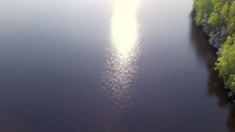 Wunderschöne-Luftaufnahmen-Eines-Abgelegenen-Waldsees-Mit-Entspannenden-Sonnenstrahlen,-Die-Von-Der-Oberfläche-Des-Sees-In-Finnland-Reflektiert-Werden