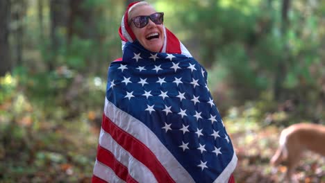 Blonde-Frau,-Die-Hinter-Sich-Mit-Lächelndem-Ausdruck-Eine-Amerikanische-Flagge-Hisst-Und-Die-Flagge-Um-Ihren-Kopf-Und-Körper-Wickelt