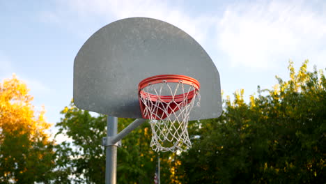 Sliding-left-shot-of-a-basketball-hoop-with-a-metal-backboard,-orange-rim-and-net-in-an-empty-park-court-at-sunrise