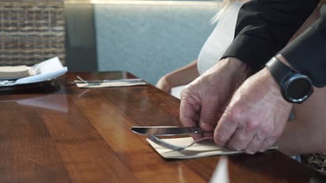 Camarero-Masculino-Poniendo-Cubiertos-En-Una-Mesa-De-Madera-Mientras-Una-Joven-Rubia-Mira-El-Menú-En-Un-Restaurante-De-Lujo---De-Cerca