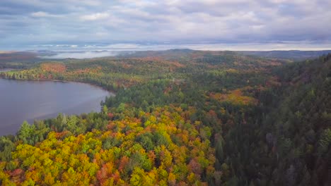 Aerial-Daytime-Wide-Shot-Flying-Over-Fall-Forest-Colors-And-Calm-Misty-Lakes-Panning-Right-To-Reveal-Sun-And-Huge-Tall-Rocky-Cliff-Ridge-in-Kawarthas-Ontario-Canada