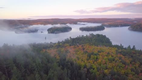 Weitwinkelaufnahme-Des-Sonnenaufgangs-Aus-Der-Luft,-Die-über-Herbstwaldfarben-Auf-Nebligen-Felsrücken-Fliegt,-Schwenkt-Direkt-In-Richtung-Des-Nebligen-Sees-Mit-Nebelbedeckten-Kieferninseln-Und-Rosa-Wolken-In-Kawarthas,-Ontario,-Kanada