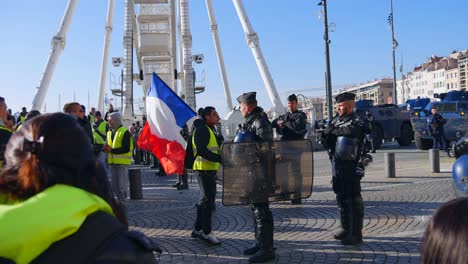 Yellow-jacket-demonstrator-with-a-french-flag-and-sun-glasses-walks-towards-a-police-officer-in-full-riot-gear-and-points-a-finger-at-him