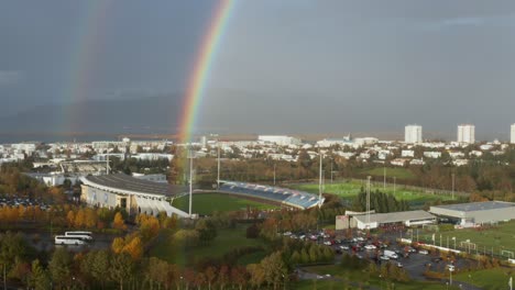 Colorful-rainbow-above-football-stadium-in-capital-city-Reykjavik,-aerial