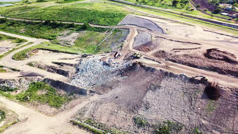 Drone-circling-in-arc-around-large-pile-of-rubbish-at-landfill-site,-surrounded-by-green-vegetation