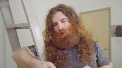 Long-auburn-hair-and-beard-clad-man-chatting-and-smiling-with-customer-out-of-frame-indoors-holding-on-to-ladder-and-tools-scraper-talking-happy-about-work