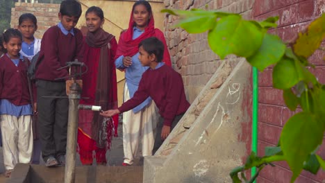 Un-Grupo-De-Escolares-Pakistaníes-De-La-Zona-Rural-Que-Usan-Una-Bomba-Manual,-En-La-Temporada-De-Invierno,-Usan-Suéteres-Y-Chaquetas,-Son-Divertidos-Y-Sonrientes,-Una-Pared-De-Ladrillos-Rojos-Y-Una-Antigua-Puerta-De-La-Escuela