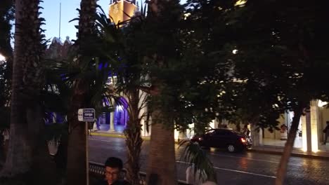 Dolly-shot-to-the-left-through-the-trees-of-the-Municipal-building-at-dusk-with-its-blue-lights-next-to-the-plaza-grande-in-Merida,-Mexico