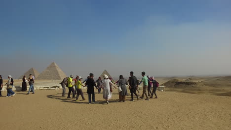 Cámara-Siguiendo-A-Los-Turistas-Que-Bailan-En-Grupo-Y-Tomándose-De-La-Mano-Para-Las-Cámaras-Frente-Al-Complejo-Piramidal-De-Giza