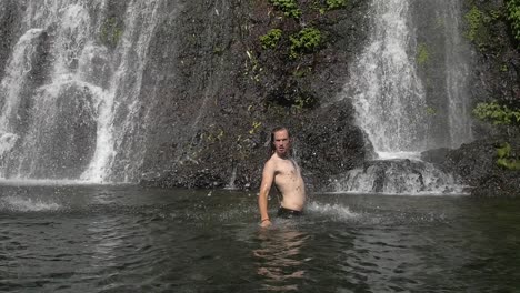 Slow-motion-of-a-funny-man-with-long-hair-in-a-waterfall-emerging-from-pool-water---flicking-his-hair-back-as-he-falls-slowly-back-into-water