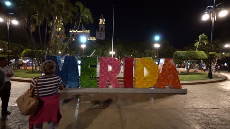 Empujando-El-Cartel-De-Merida-Para-Que-La-Gente-Se-Tome-Selfies-Frente-A-La-Catedral-De-San-Ildefonso-Al-Fondo,-Al-Atardecer