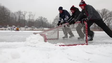 Tres-Jóvenes-Que-Usan-Su-Portería-De-Hockey-Para-Empujar-La-Nieve-Del-Campo-De-Hockey-De-Su-Estanque-Hacia-La-Cámara