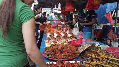 Muchas-Personas-De-Pie-Y-Comprando-Comida-Al-Vendedor-Con-Un-Montón-De-Diferentes-Tipos-De-Carne-En-La-Mesa-Con-Bolsas-De-Plástico-Colgadas-Y-Bombillas