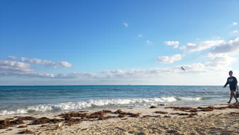 Gente-Caminando-Por-La-Playa-Con-Parapente-En-El-Horizonte-Con-Olas-Rompiendo-En-México-En-Un-Día-Soleado