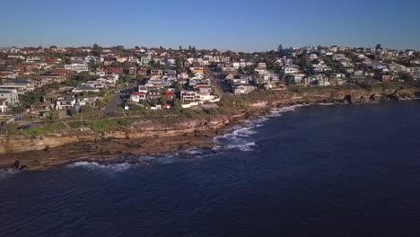 Beautiful-reverse-drone-shot-of-houses-near-the-beach-and-cliff-with-view-of-pacific-ocean-waves-crashes-the-rocks