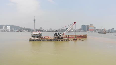 Niedriger-Drohnen-LKW-Schuss,-Der-An-Einem-Trichterkahn-Vorbeifährt,-Und-Einem-Kobelco-Kobe-Stahlgreifbagger-Mit-Der-Skyline-Von-Macau-Im-Hintergrund