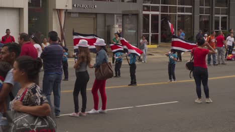 Erwachsene-Beobachten,-Wie-Kleine-Kinder-Während-Der-Parade-Zum-Unabhängigkeitstag-Von-Costa-Rica-Mit-Fahnen-Marschieren