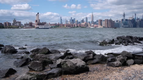 Panorámica-Lenta-A-Través-Del-Horizonte-De-La-Ciudad-De-Nueva-York-Con-Olas-Rompiendo-En-La-Playa-Desde-El-Río