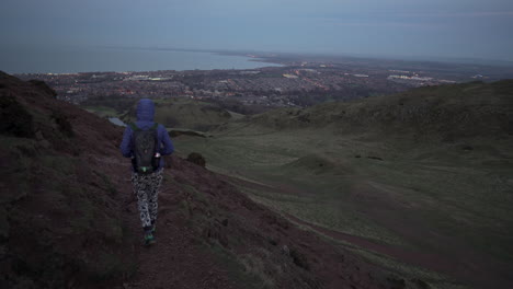 Follow-tracking-shot-of-girl-walking-down-the-Arthurs-seat-mountain-on-the-hiking-trail-in-evening,-dusk-with-city-of-Edinburgh-and-Atlantic-Ocean-in-the-background