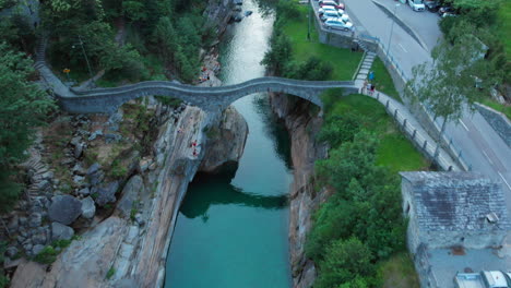 Wide-revealing-shot-of-a-roman-bridge-at-Verzasca-valley,-two-people-jump-from-the-cliff