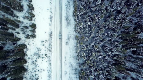 Aerial-view-looking-down-as-a-car-drives-along-a-winter-road-through-pine-forest